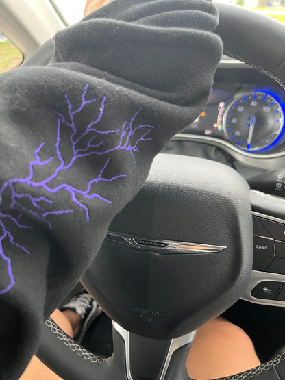 Hand on the Steering wheel of a car with a hand wearing a black sweater with purple lightning on the sleeve.